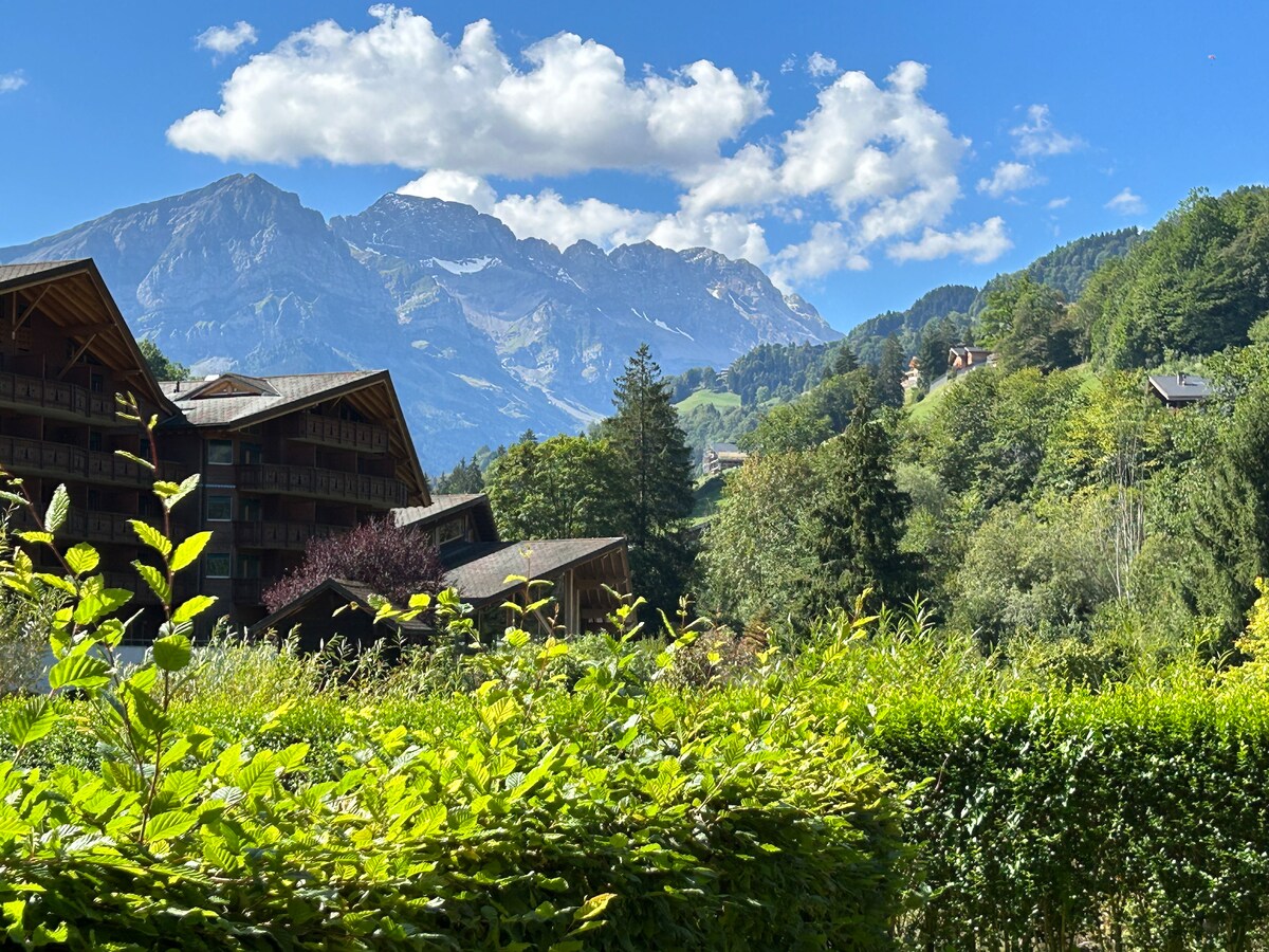 A panoramic view captures the majestic Dents Du Midi mountains under a partly cloudy sky. Green foliage frames the scene, showcasing the natural beauty of the surrounding area with its varied textures and colors. The peaceful landscape reflects the alpine environment.