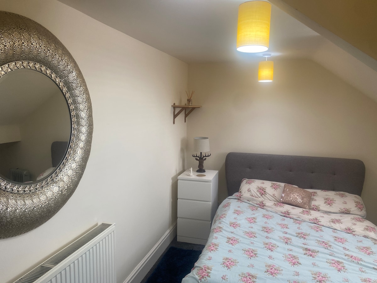 A cozy bedroom features a comfortable double bed dressed in floral bedding. A modern mirror hangs on the wall, reflecting soft light from ceiling lamps. A small bedside table and a white storage unit add functionality to the space, complemented by neutral-colored walls.