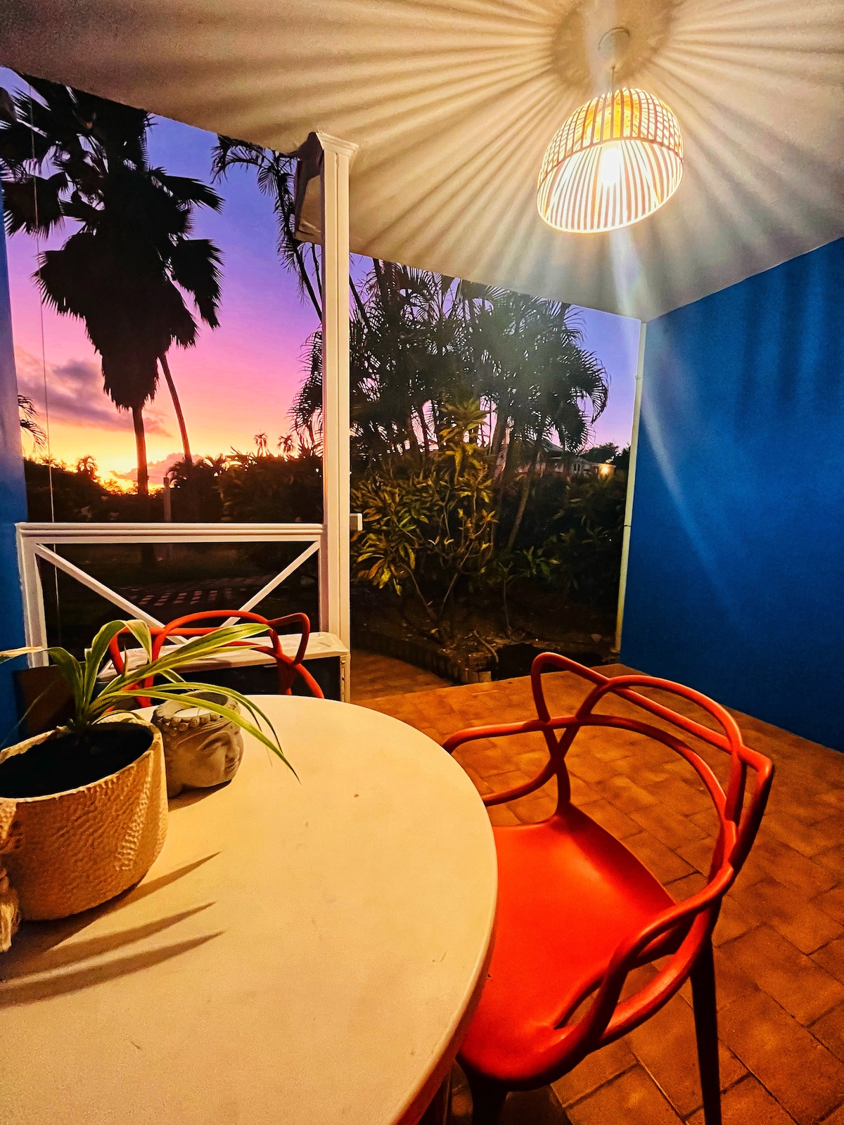 An outdoor seating area features a round table surrounded by colorful chairs. A potted plant adds a touch of greenery. The backdrop showcases a vibrant sunset, with palm trees silhouetted against the colorful sky, creating a serene and inviting atmosphere.