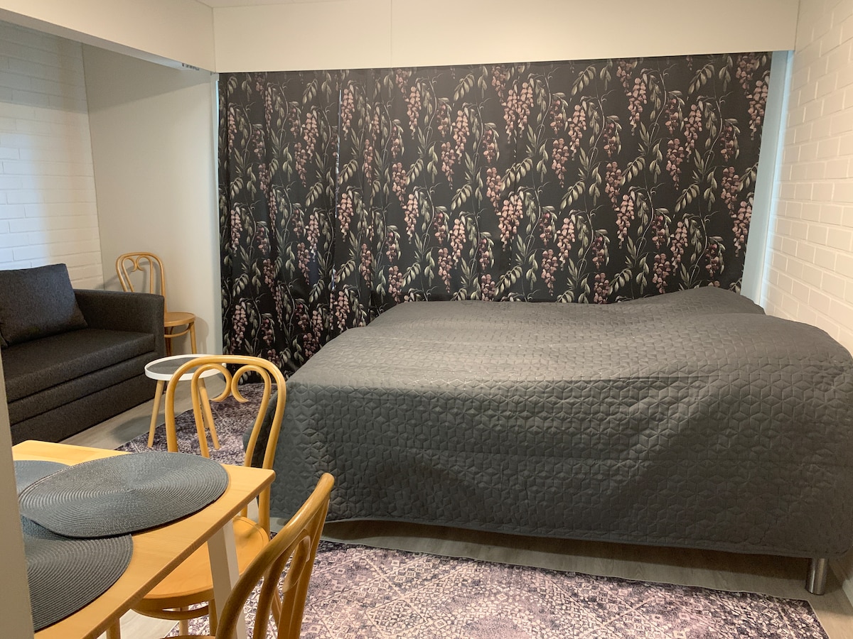 A spacious sleeping area features a large bed covered by a dark quilt. A textured area rug lies underneath. To the side, a cozy sofa offers relaxation space. Two wooden chairs surround a simple dining table, all set against a floral accent wall.