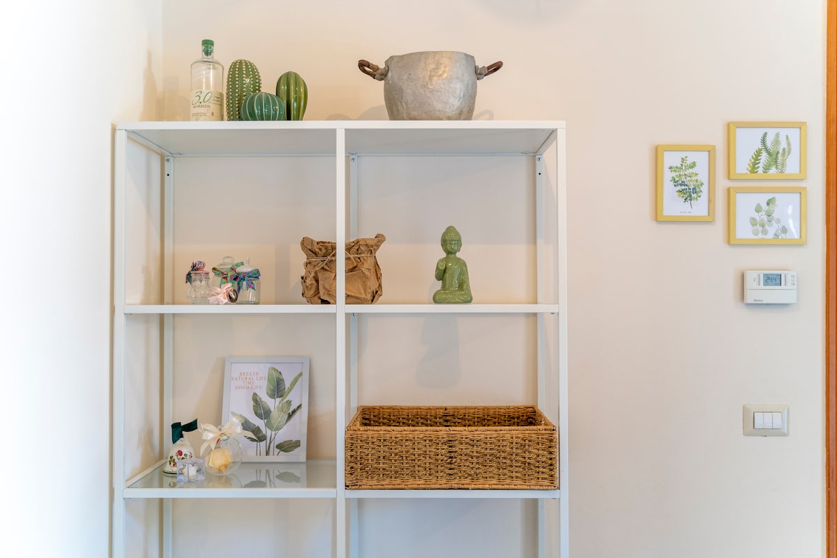 A modern white shelving unit is displayed against a wall, featuring various decorative items. Potted plants and a metallic bowl are placed on the top shelf. The lower shelves contain a woven basket and a framed botanical print alongside a small statue.