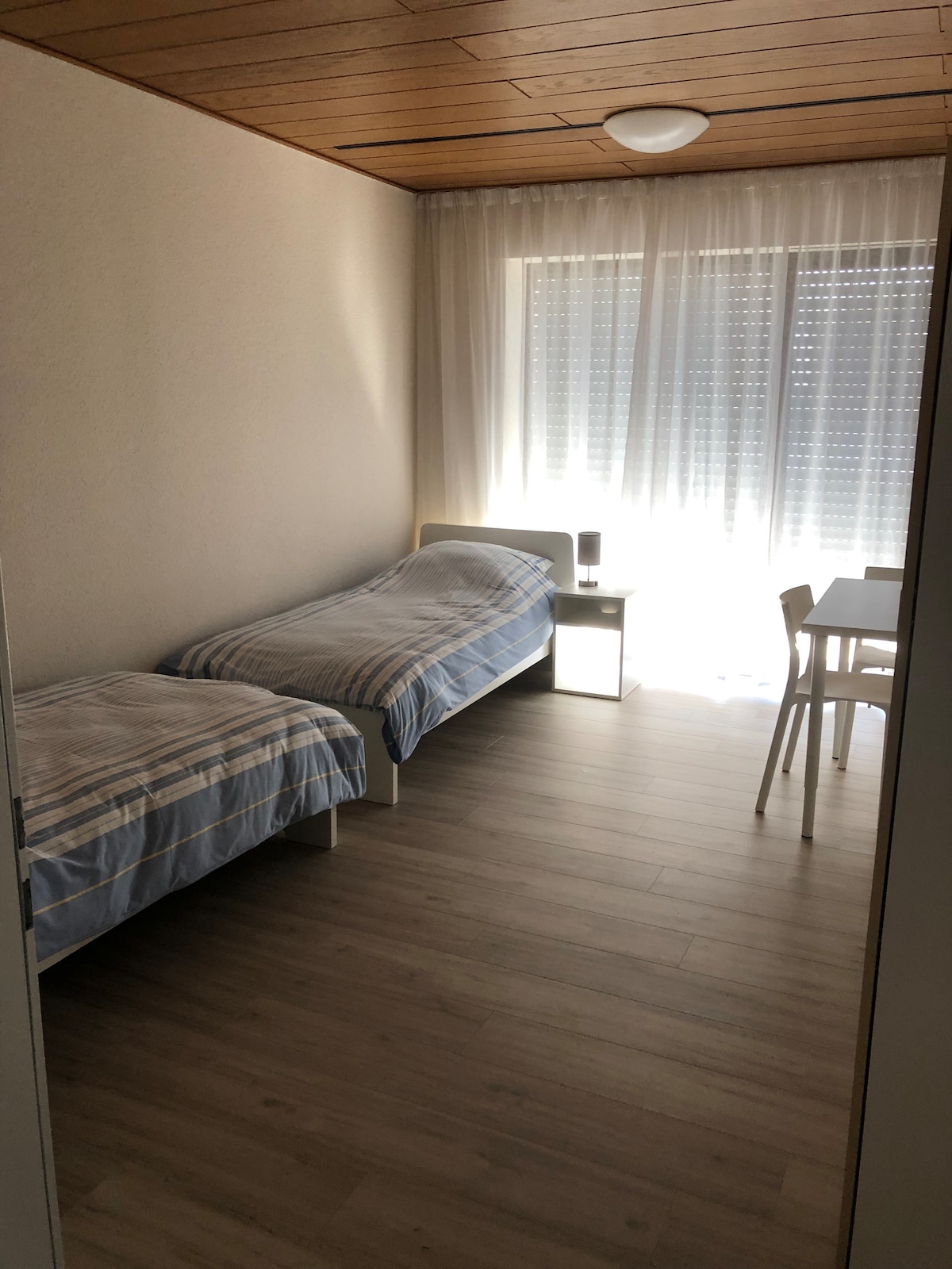 A bright bedroom is presented with two twin beds, each dressed in light blue striped linens. Natural light floods the space through sheer curtains covering large windows. A small table and chair set is positioned in the corner, contributing to the room's functional layout.