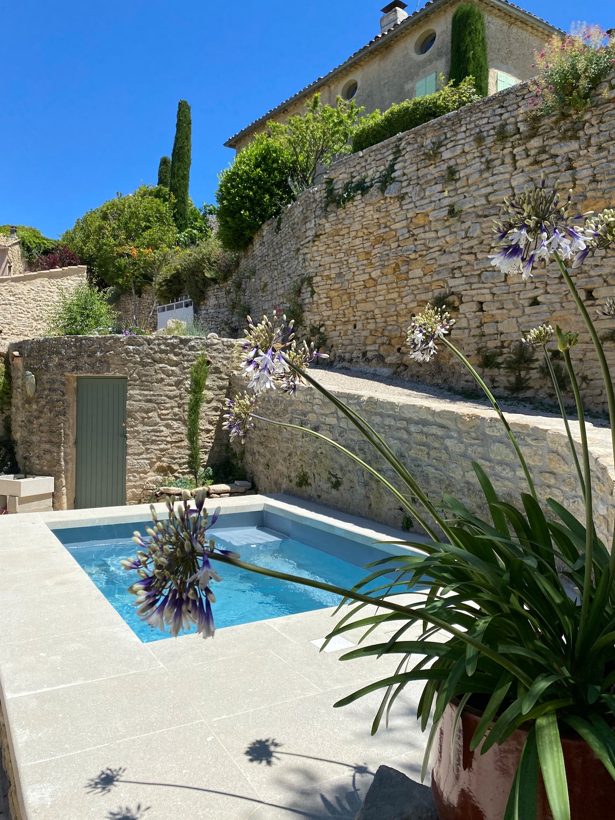 The image displays a private stone terraced area featuring a rectangular heated pool surrounded by lush green plants and flowering features. The serene stone walls and steps create an inviting backdrop against a clear blue sky, enhancing the sense of tranquility.