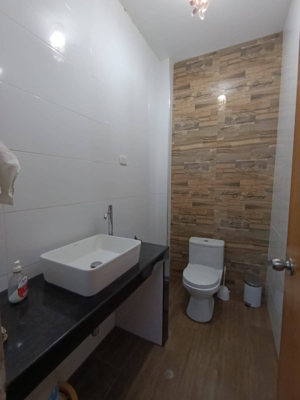 A modern bathroom features a compact design with a rectangular sink atop a sleek black countertop. The wall is partially covered with decorative wood-patterned tiles, while the rest presents a glossy white finish. A toilet is positioned discreetly, and light fixtures offer bright illumination.