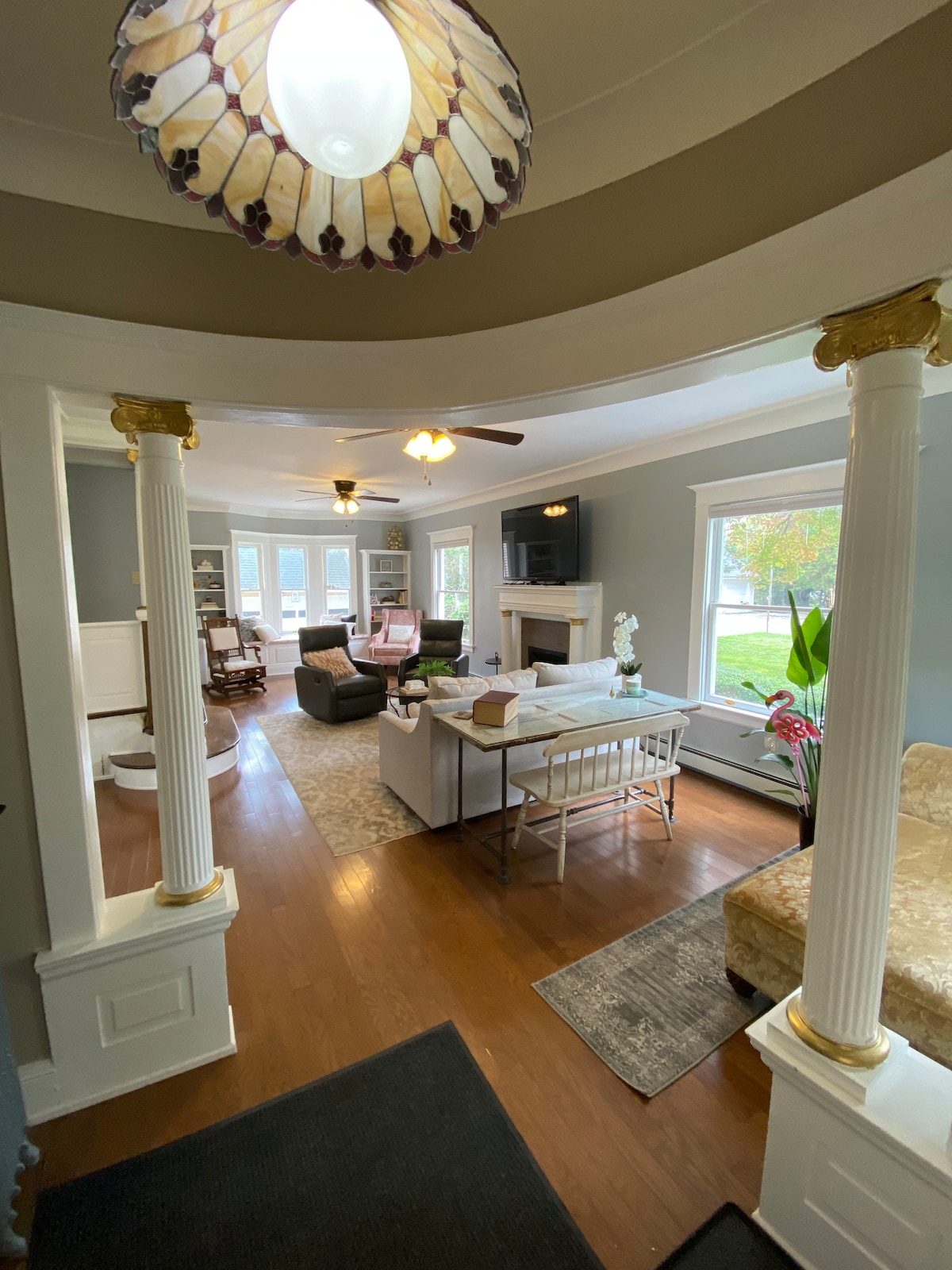 A welcoming living area is framed by elegant columns, featuring a cozy seating arrangement with a mix of soft furniture. Natural light fills the space through multiple windows, highlighting a decorative area rug on polished hardwood floors.