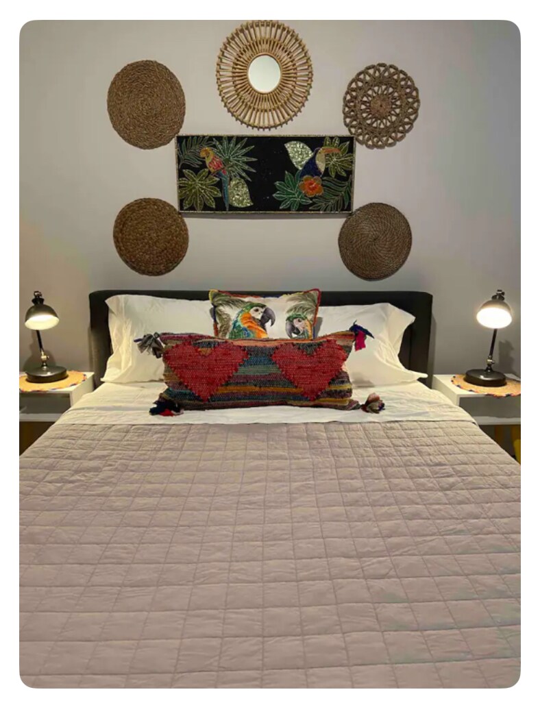 A cozy bedroom is showcased with a spacious bed dressed in a light quilt. Decorative pillows featuring colorful designs are positioned at the head of the bed. Wall art made of woven materials and a central tapestry with tropical patterns accentuate the serene atmosphere.