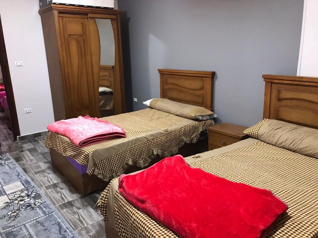 Two twin beds are arranged in a comfortable layout, each dressed with distinct blankets. A wooden wardrobe with a mirrored door adds storage and reflects the room's design. The floor is covered with patterned tiles, while the walls are painted in a neutral tone.
