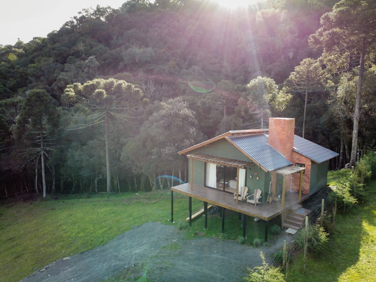 A cabin is nestled among lush greenery, featuring a wooden deck that extends outward. Two chairs are positioned for outdoor relaxation, while a chimney adds a touch of character. Natural light filters through the trees, highlighting the cabin's rustic charm.