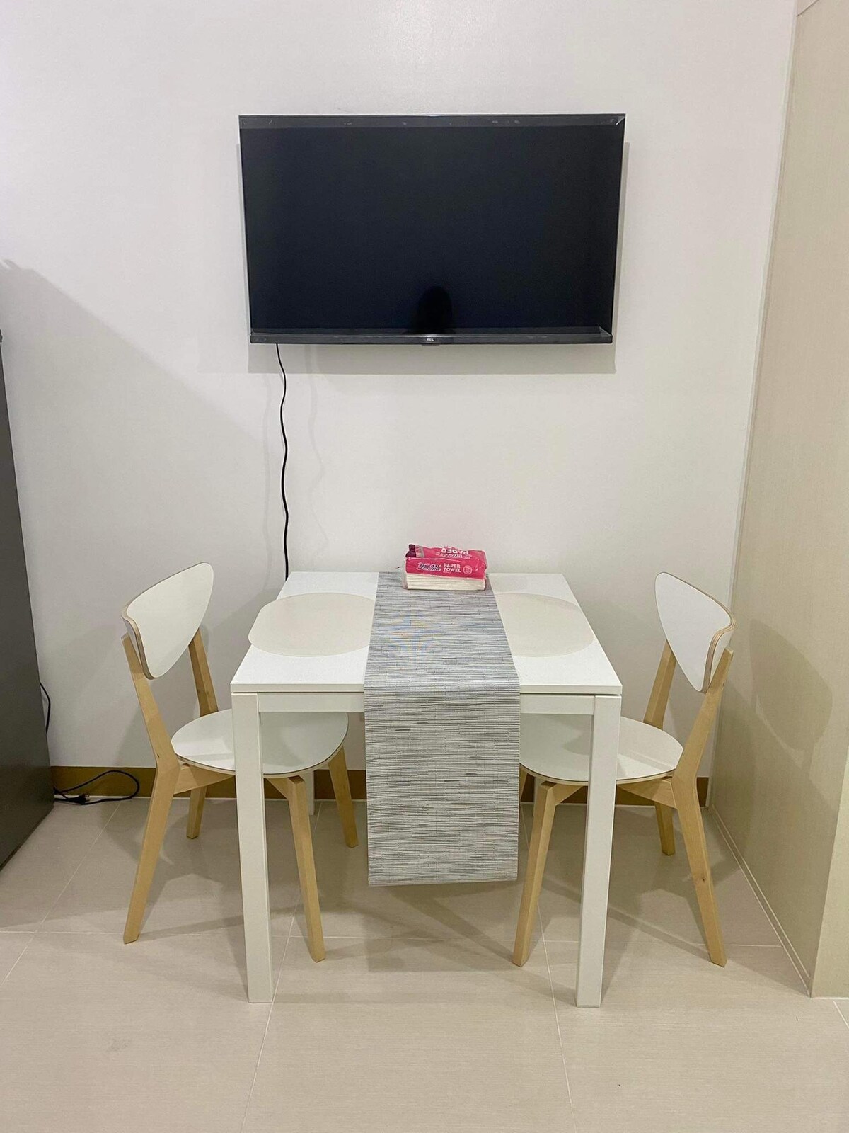 A small dining area is featured, consisting of a white table and three light wooden chairs. A simple table runner lies across the table. A flat-screen television is mounted on the wall above the table, creating a functional space for meals or relaxation.