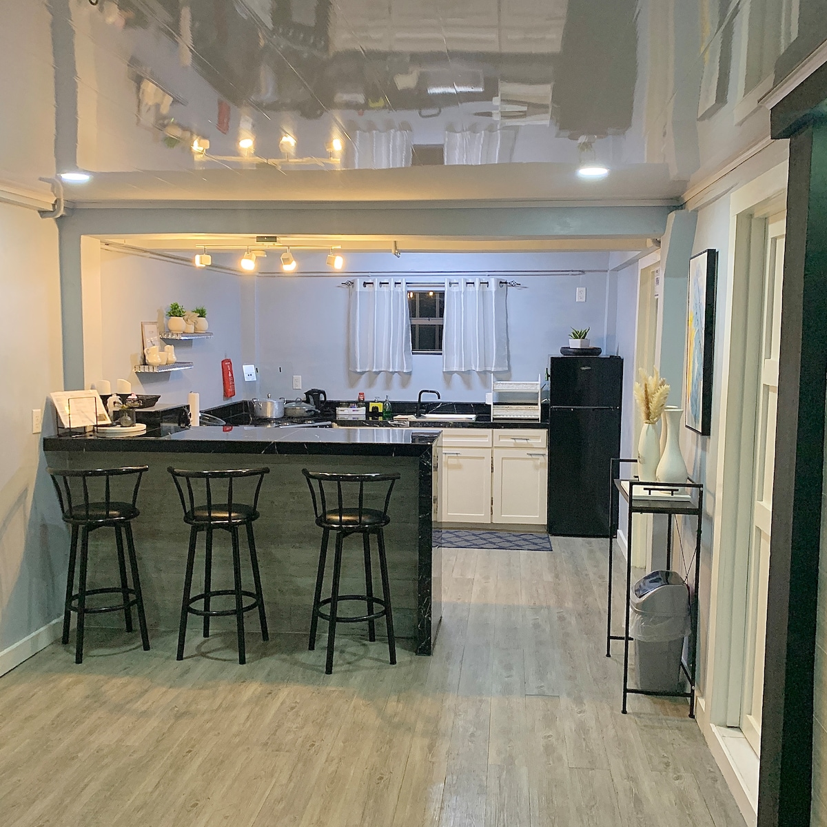 A modern kitchen features a central island with three black bar stools. Countertops are lined with appliances, and cabinets offer ample storage. Natural light enters through a window, while the walls are painted in soft tones, creating a bright and functional space.