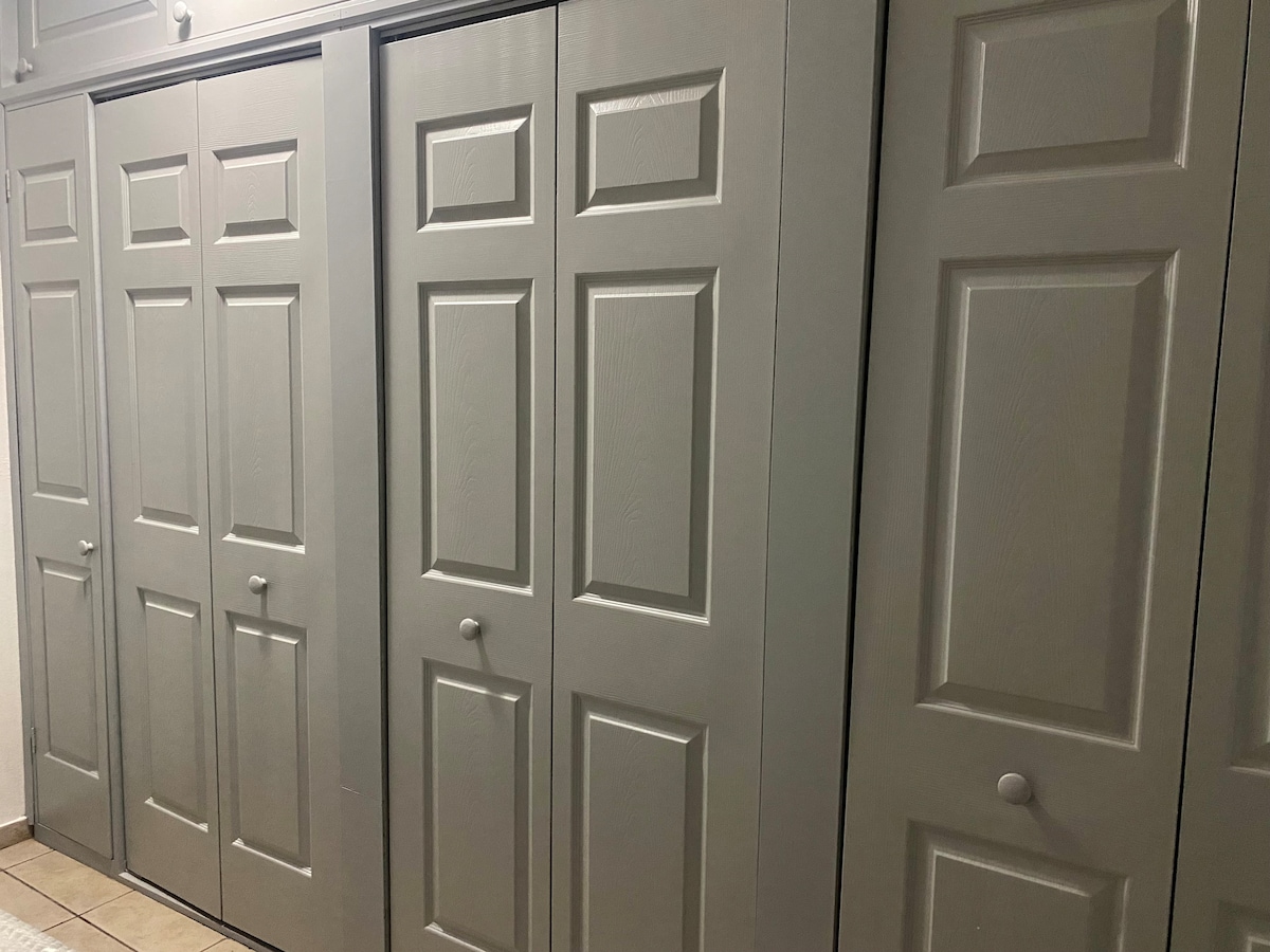 Shuttered storage space is showcased, featuring multiple grey doors with flat panel designs. The doors are set within a light-colored wall, providing ample storage options for guests. A tiled floor is visible beneath, contributing to a neutral and functional ambiance.