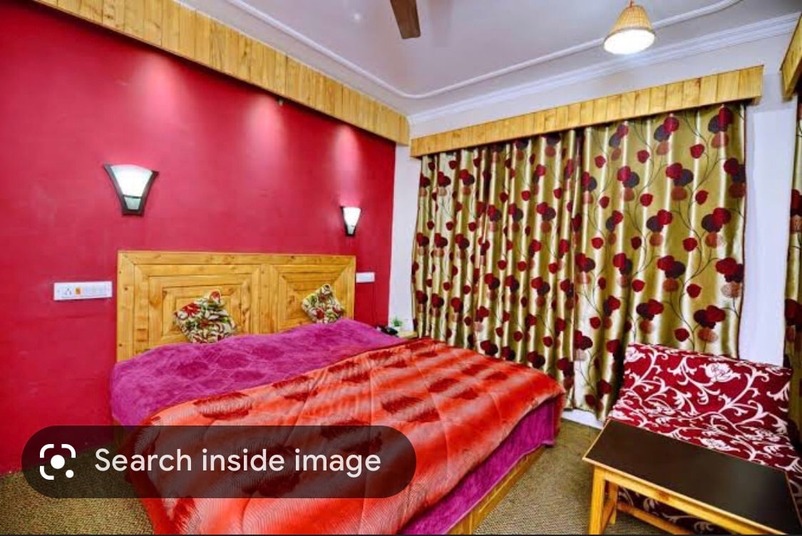 A warm-toned bedroom features a spacious bed adorned with a vibrant blanket. Floral-patterned curtains frame the window, complemented by wooden accents. Soft lighting from wall sconces enhances the cozy atmosphere. A small table and chair offer additional seating in the inviting space.