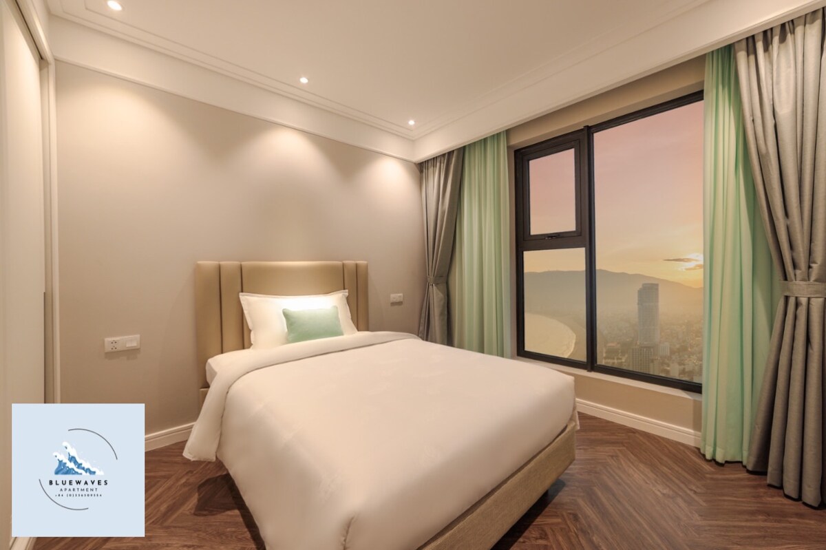 A spacious bedroom is presented with a soft-colored wall and ample natural light pouring in from a large window. A comfortable bed with a light-colored headboard is dressed in crisp white linens, complemented by a decorative pillow. Floor-to-ceiling curtains frame the window.