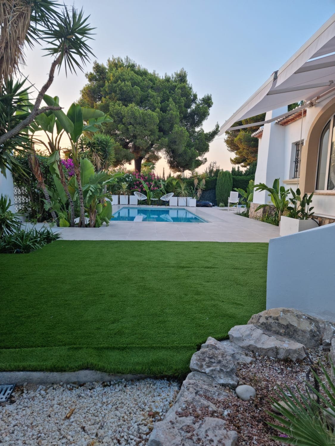 A neat outdoor area features manicured green grass and tropical plants. A swimming pool is visible in the distance, surrounded by palm trees and flowering shrubs. The exterior of the villa is partially seen on the right, with a spacious terrace providing a serene atmosphere.