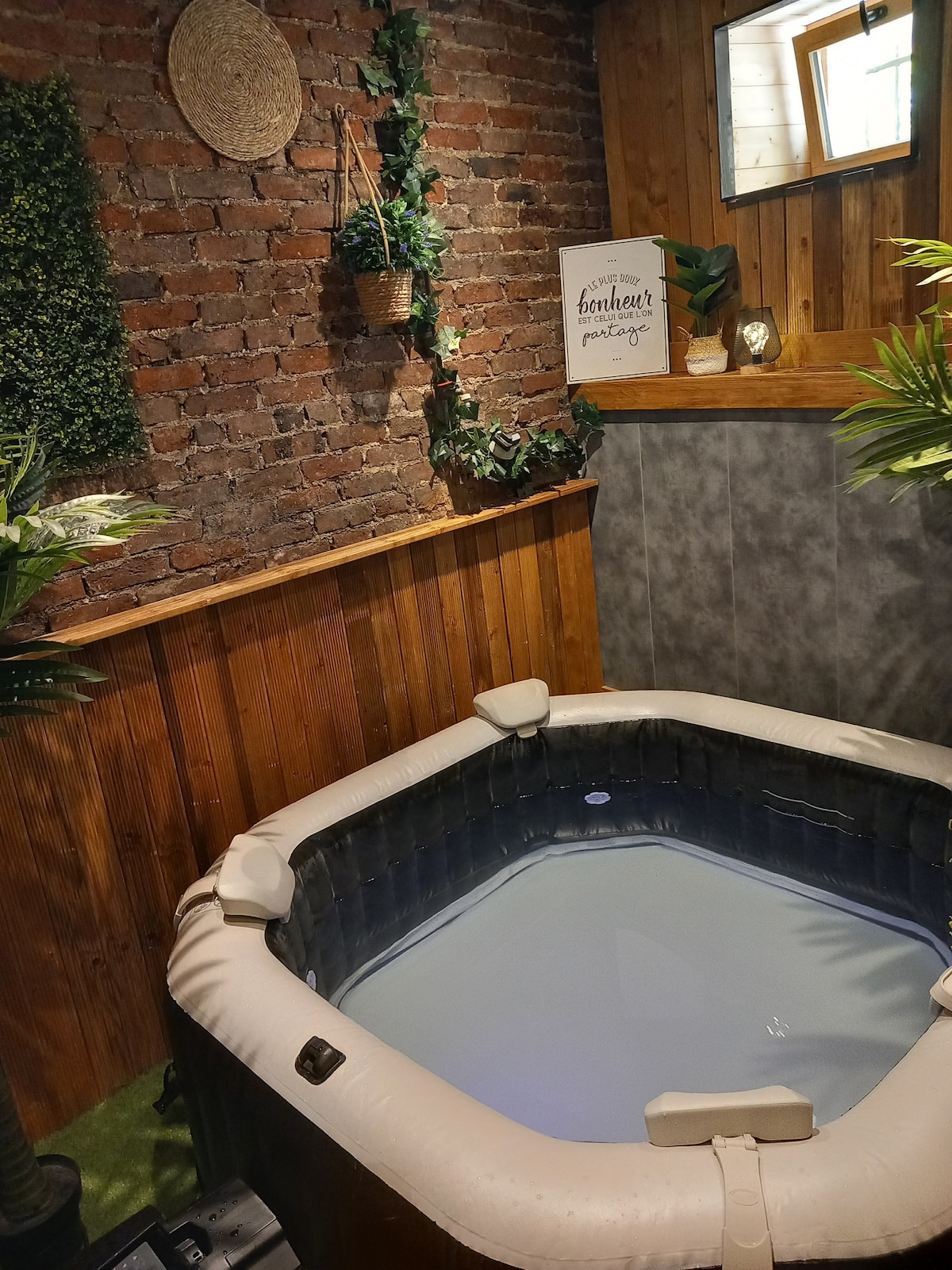 A cozy jacuzzi is set within a rustic wooden enclosure, accented by exposed brick walls and greenery. The calming water reflects soft lighting, creating a serene atmosphere. A mirror and decorative elements enhance the space, lending a touch of comfort and relaxation.