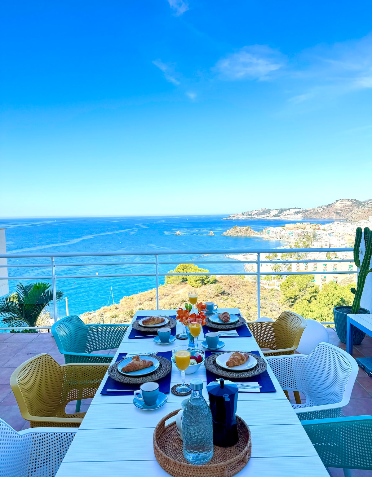 A dining table is set with colorful tableware, including plates and glasses, overlooking a bright blue sea. Delicate pastries are arranged alongside fresh juice and water. The backdrop features a clear sky and coastal landscape, highlighting the inviting outdoor dining experience.