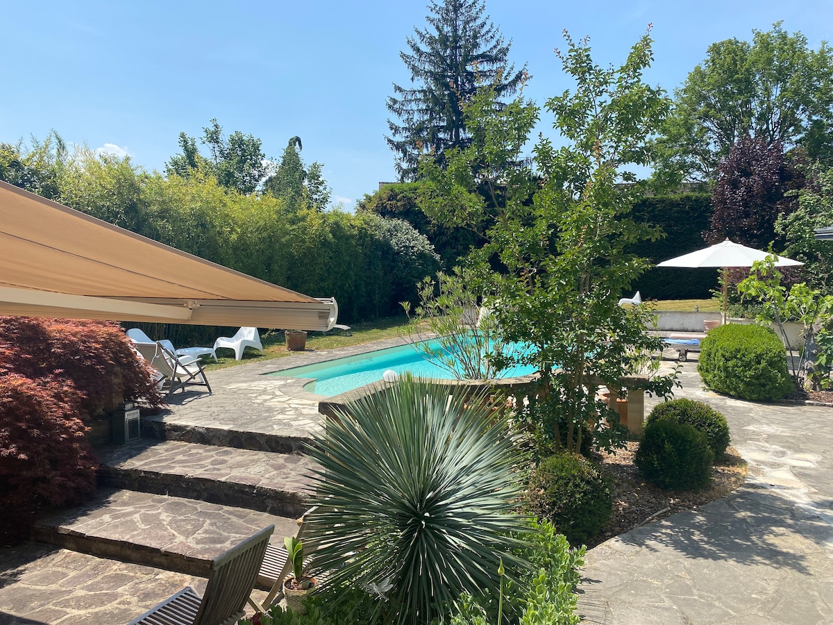 A serene outdoor space is highlighted, featuring a landscaped garden with various greenery, including a palm-like plant and shrubs. An inviting pool is centrally located, surrounded by sun loungers and umbrellas for shade. A sun awning provides additional coverage over the patio area.