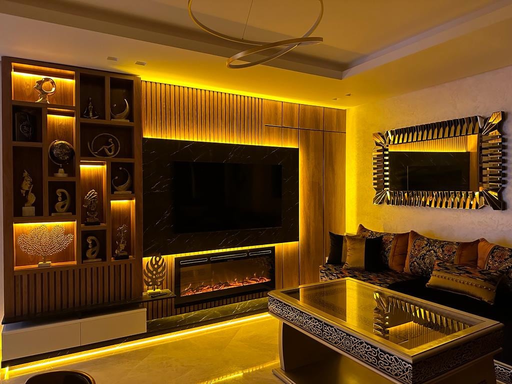 A modern living room features warm ambient lighting that highlights textured wooden panels and decorative shelving. A sleek television is mounted on the wall above a contemporary electric fireplace. A stylish coffee table sits in the center, surrounded by comfortable seating adorned with decorative cushions.