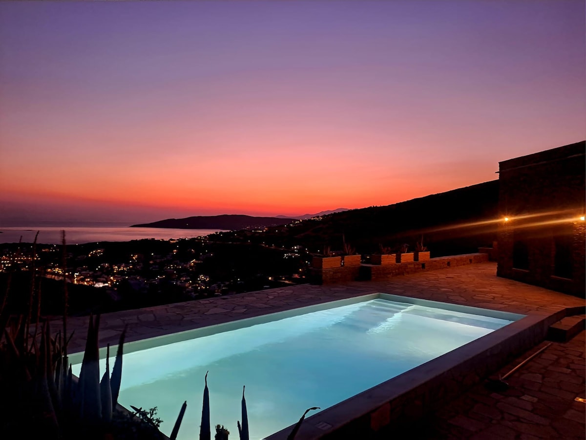 An inviting swimming pool reflects the soft hues of twilight, surrounded by serene landscapes. Hills and the Aegean Sea are visible in the distance, where the horizon showcases a gradient of warm colors as the sun sets. Soft lighting enhances the serene ambiance.