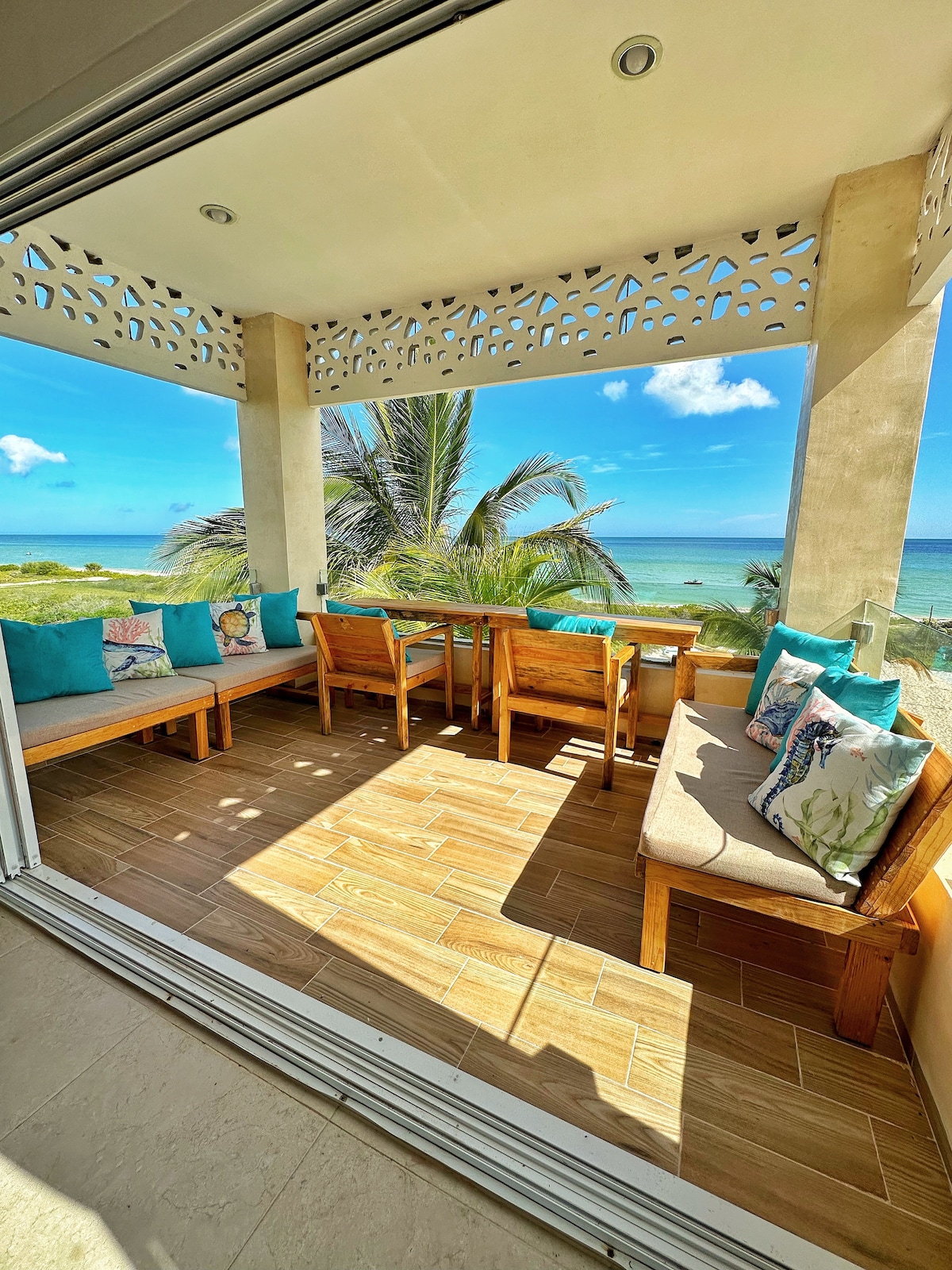 A charming terrace showcases natural light flooding in, enhanced by comfortable seating adorned with colorful cushions. The view features a vast seascape, accented by swaying palm trees under a clear blue sky.