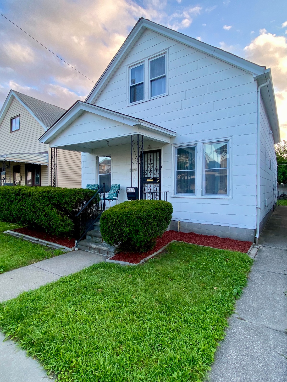 A two-story house features a white exterior with a sloped roof and a small front porch, complemented by decorative railings. Two large windows allow natural light into the home. Well-maintained shrubs and manicured grass enhance the inviting exterior.