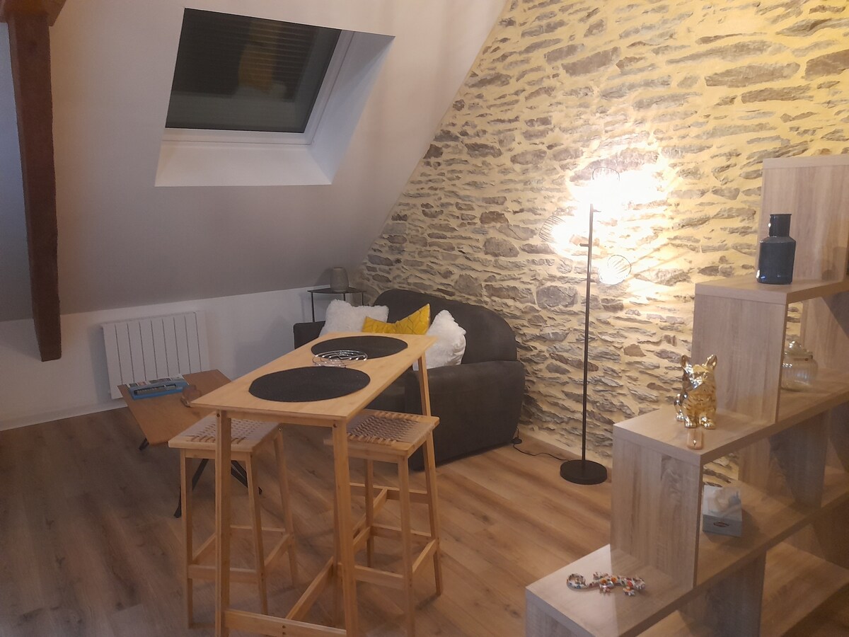A cozy living space features a sloped ceiling and a combination of stone and wood elements. A comfortable sofa is positioned next to a tall floor lamp, while a multi-level wooden shelf displays decorative items. A small dining area is set with a round table and stools.