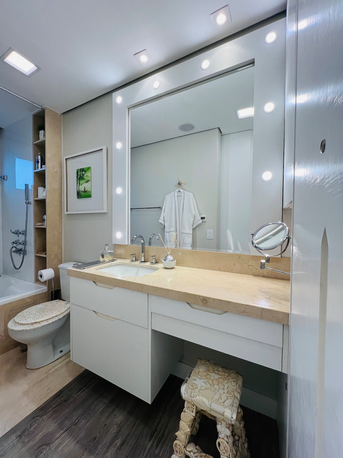 The bathroom features a dual sink vanity with a lighted mirror above, enhancing visibility. A modern shower area is visible, accompanied by a decorative artwork and a plush bathrobe hanging nearby. A small decorative stool is positioned beside the vanity.