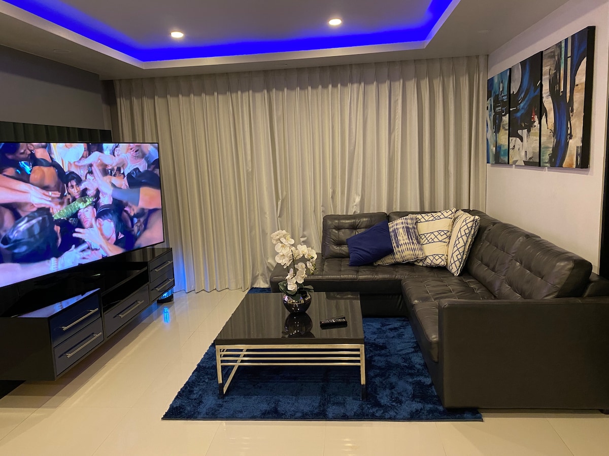 The lounge area features a modern sectional sofa adorned with decorative cushions. A large TV displays vibrant content beside a sleek black coffee table. The space is illuminated by ambient blue lighting, complemented by sheer curtains that allow natural light to filter through.