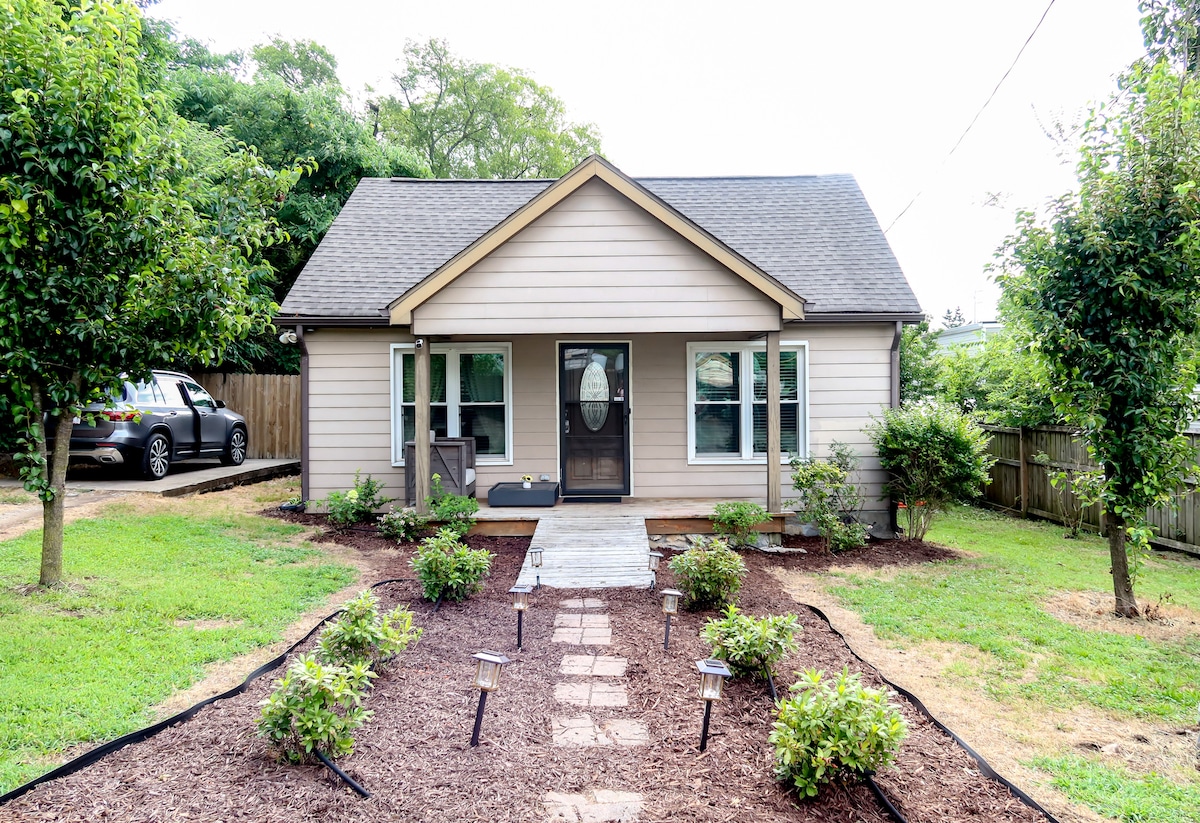 A charming cottage exterior is presented, with a light beige facade and a welcoming front porch. A stone pathway leads to the entrance, framed by neatly landscaped areas with low shrubs. Lush greenery surrounds the house, offering a tranquil outdoor space.