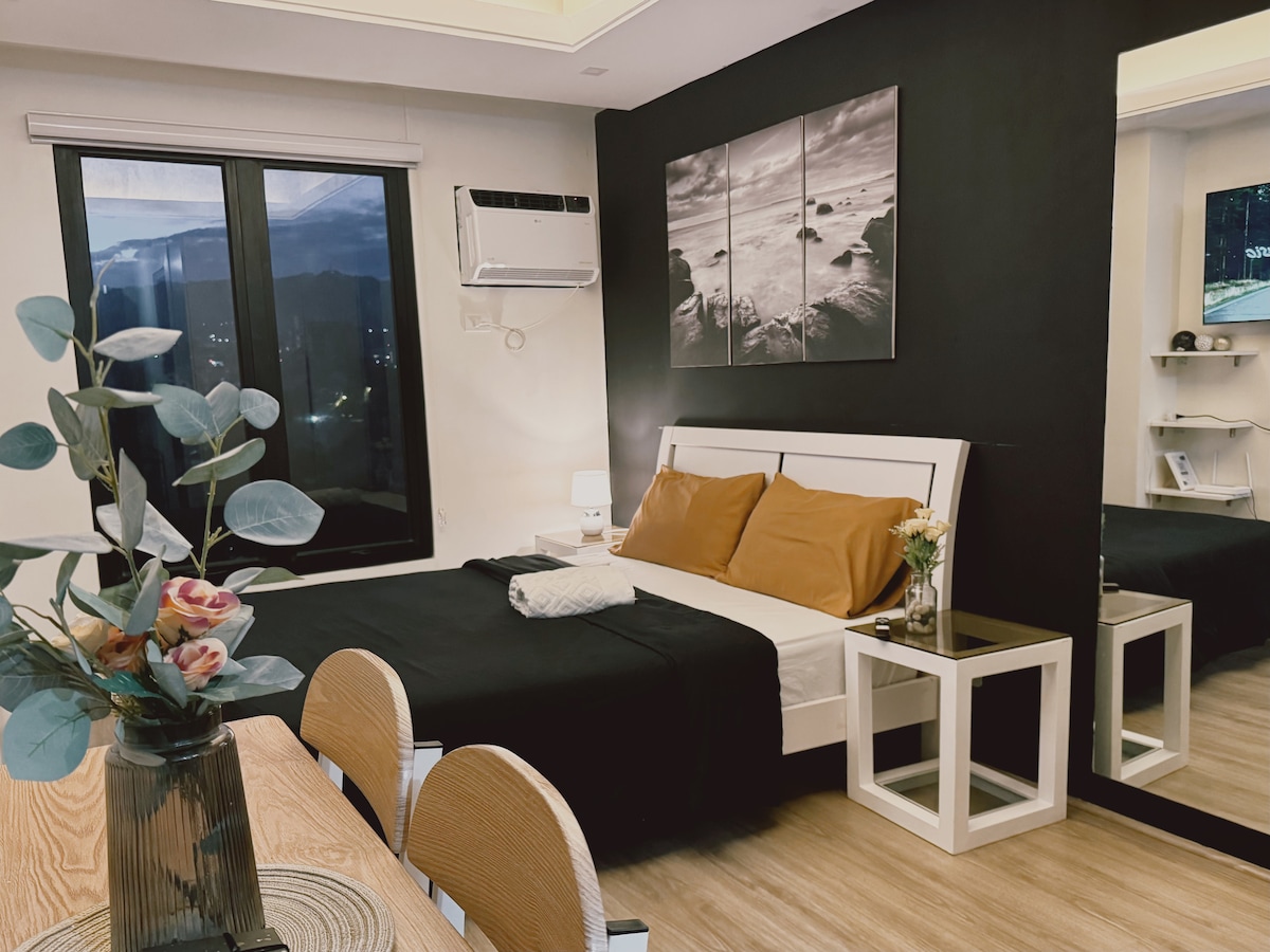 A studio apartment features a queen size bed with contrasting brown and white linens. A large window allows natural light to fill the space, complemented by an air conditioning unit mounted above. A mirror enhances the room's spacious feel, while a simple dining setup is visible.
