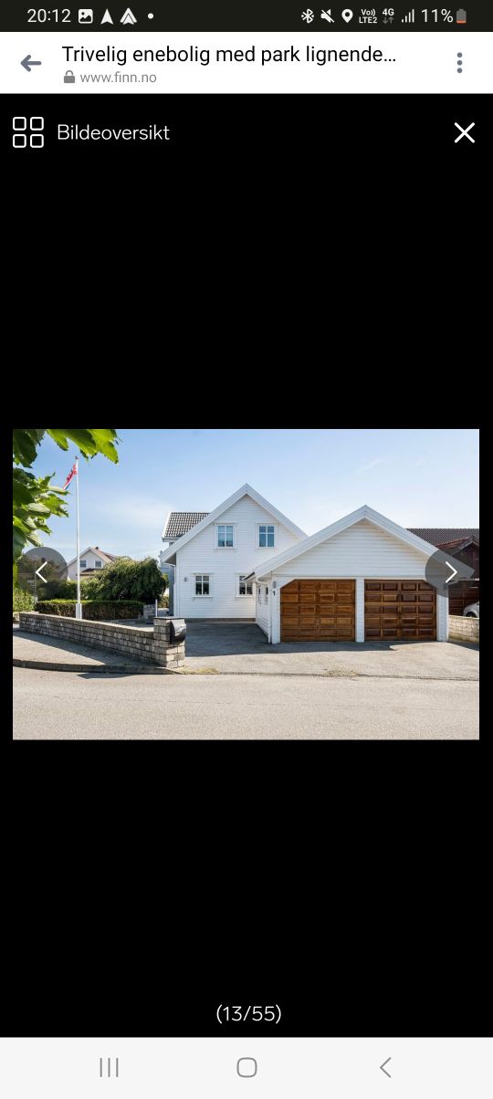 A charming house is presented, featuring a light exterior with a prominent gable roof. The entrance is accentuated by two wooden garage doors and large windows that allow natural light to fill the space. Nearby, a flagpole stands, indicating a welcoming atmosphere.