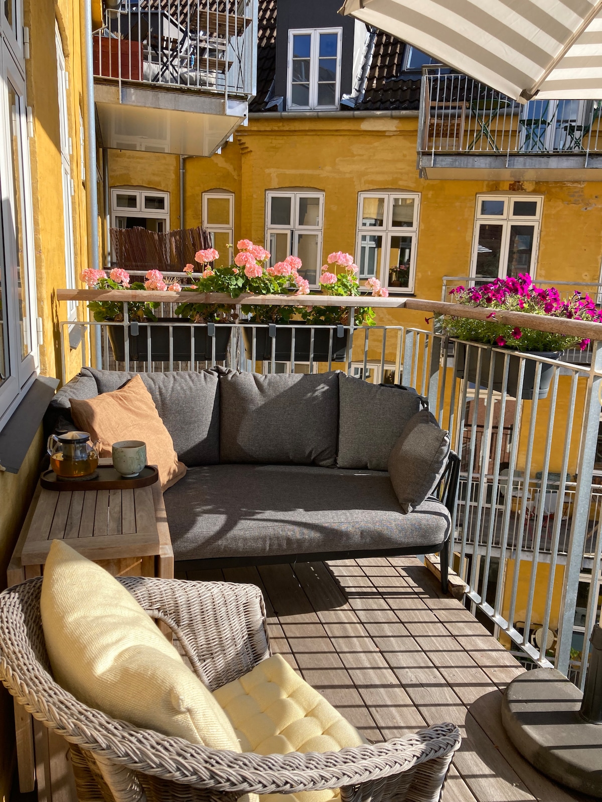 A balcony is furnished with a comfortable gray sofa and a wicker chair. Potted flowers in vibrant shades of pink add color to the space, while a wooden table holds cups and a small decorative item. Sunlight illuminates the cozy seating area.