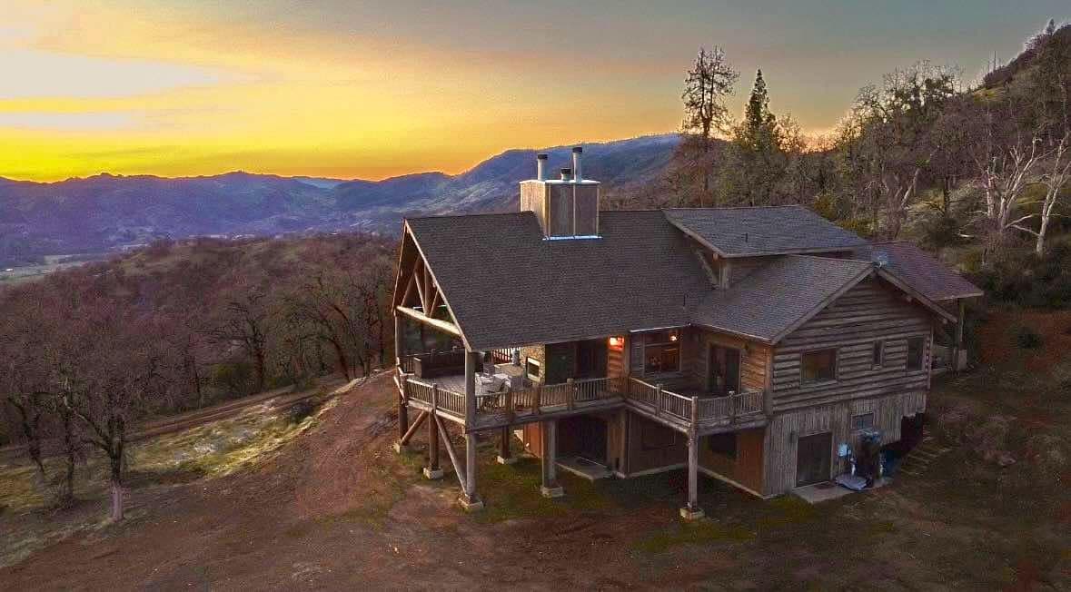A spacious lodge-style log cabin is shown, surrounded by rolling hills and stunning natural landscapes. The structure features multiple levels with a wide balcony, providing scenic views of the sunset and mountains in the distance. The earthy tones of the wood complement the serene outdoor setting.
