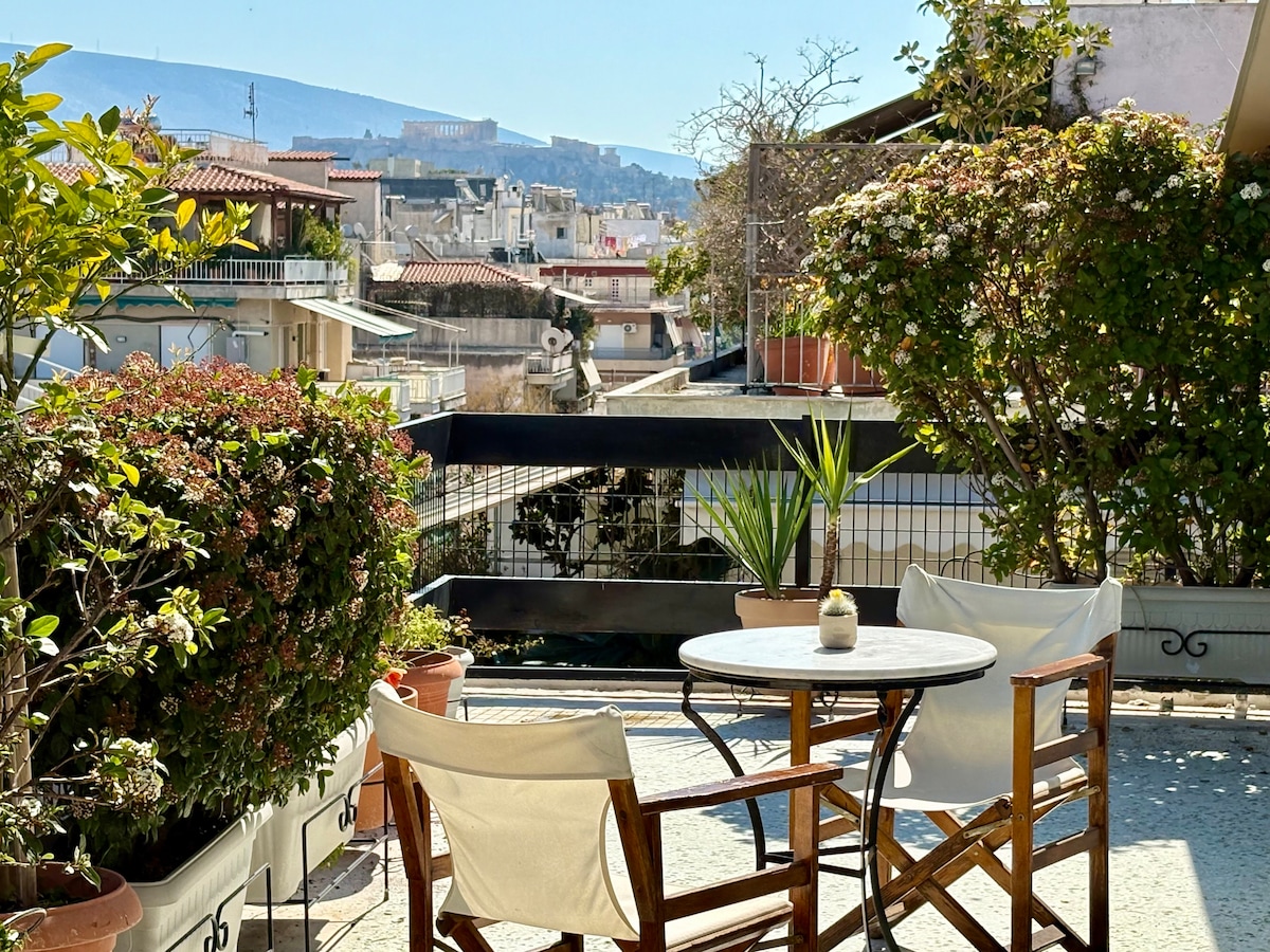 A spacious balcony is seen, adorned with greenery and potted plants. A small round table is accompanied by two wooden chairs. The view showcases neighboring buildings and distant hills, providing a glimpse of the Athenian landscape under clear blue skies.