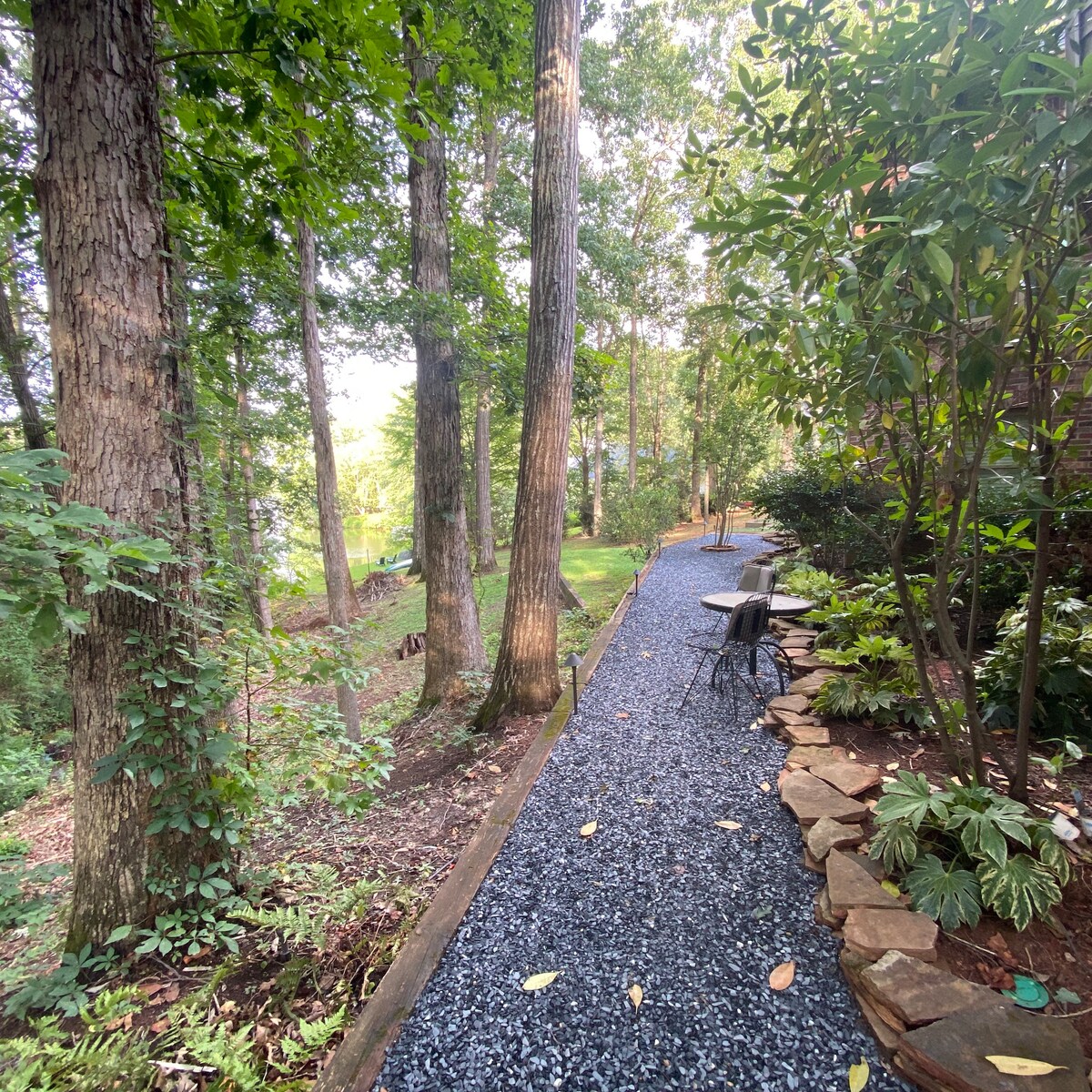 A meandering path lined with rich greenery and large trees leads to a serene outdoor space. A small seating area with two chairs is positioned next to the path, surrounded by lush plants and stones, inviting relaxation in a natural setting.