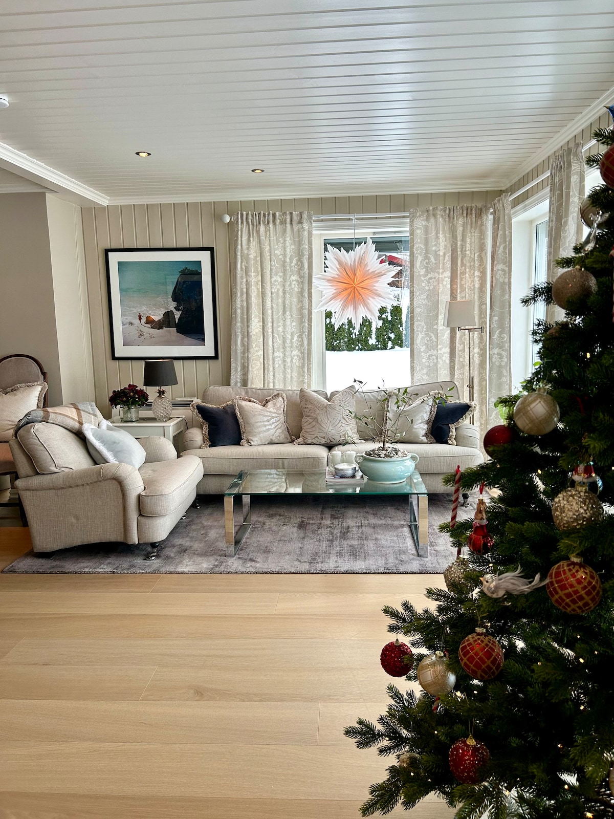 A comfortable living room features a light-colored sofa accompanied by a glass coffee table, surrounded by decorative cushions. A Christmas tree stands adorned with ornaments, while a window with sheer curtains allows natural light to illuminate the space, complementing festive decorations.