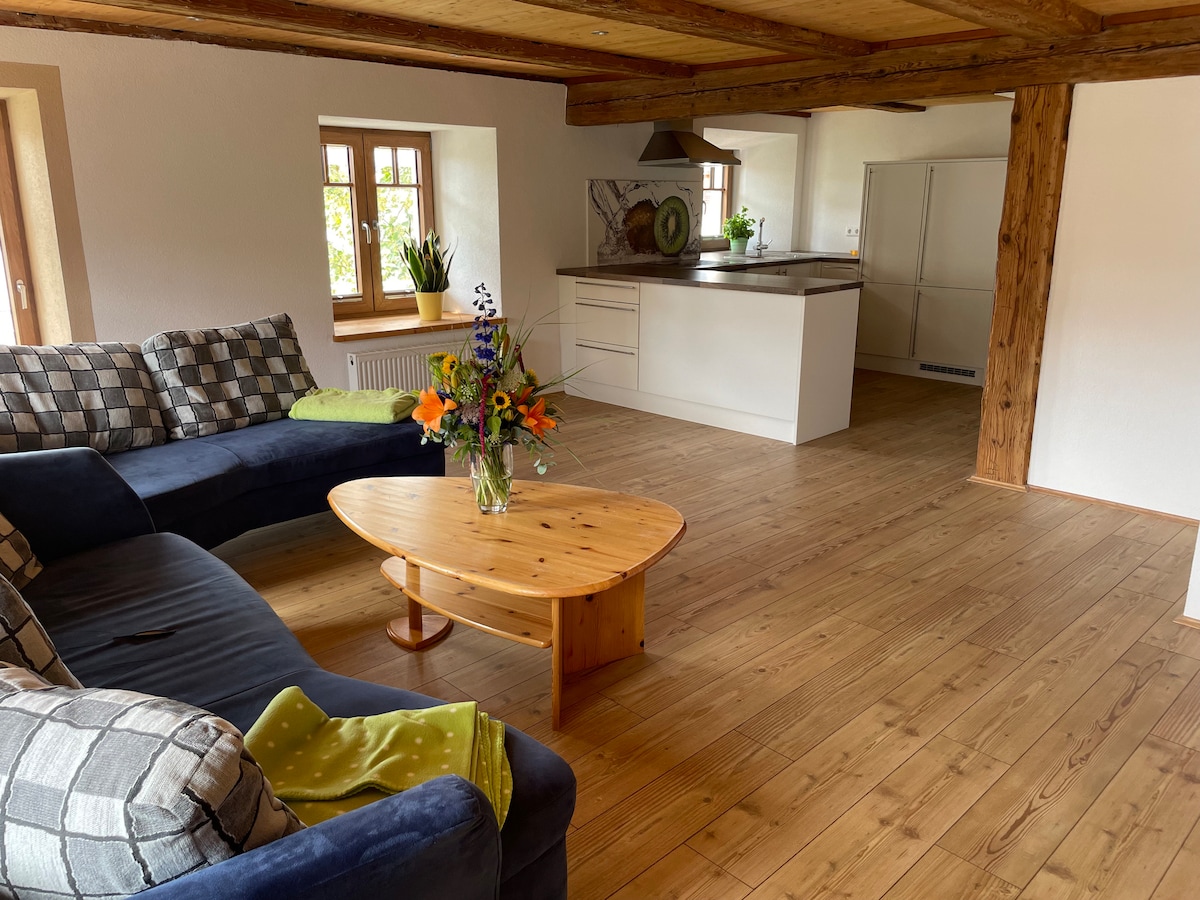 A welcoming living space features a large sectional sofa adorned with plaid and green cushions. A round wooden coffee table sits centrally, complemented by a vibrant floral arrangement. Natural light filters through the windows, highlighting the warm wooden flooring and the minimalist kitchen area in the background.