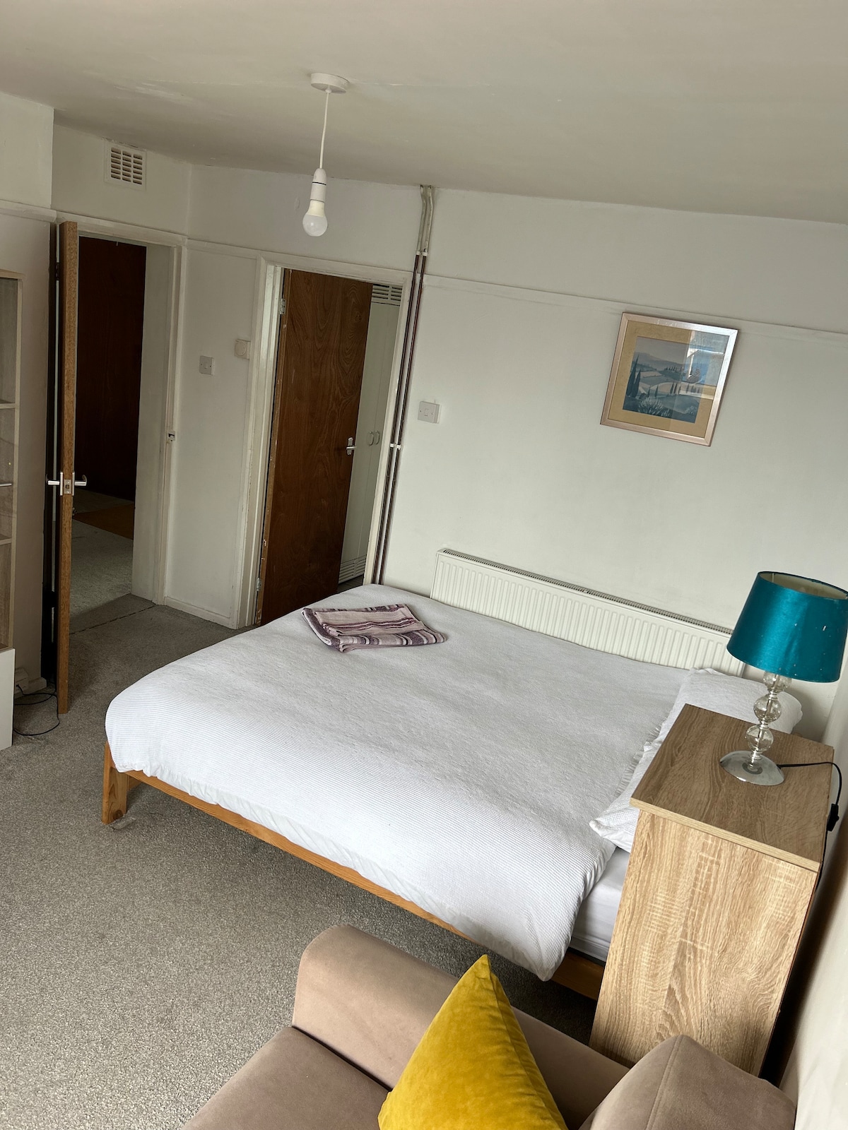 A cozy sleeping area features a spacious bed dressed in white linens. A tan-colored bedside table with a turquoise lamp complements the room's light walls. A folding door leads to a separate space, and a small sofa with a yellow cushion adds comfort.