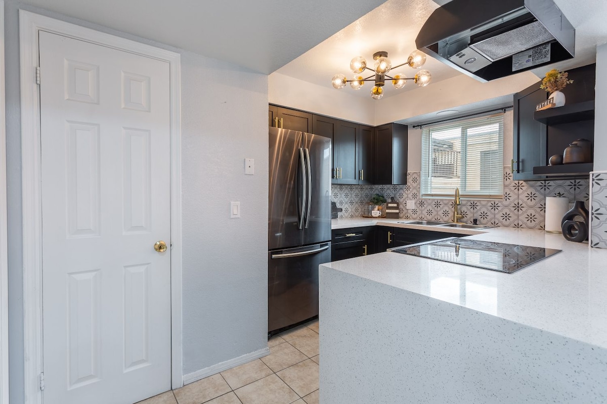 A modern kitchen is highlighted with dark cabinetry and stainless steel appliances. A large window allows natural light to illuminate the sleek countertops, complemented by geometric tile backsplashes. A door leading to another area of the home is visible, enhancing accessibility.