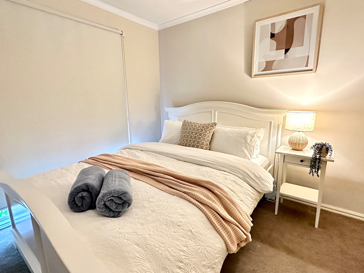 A serene bedroom features a spacious bed with crisp white linens and a soft peach throw. A bedside table holds a lamp and decorative vase, while rolled towels rest on the bed. Natural light filters through a partially closed window blind.