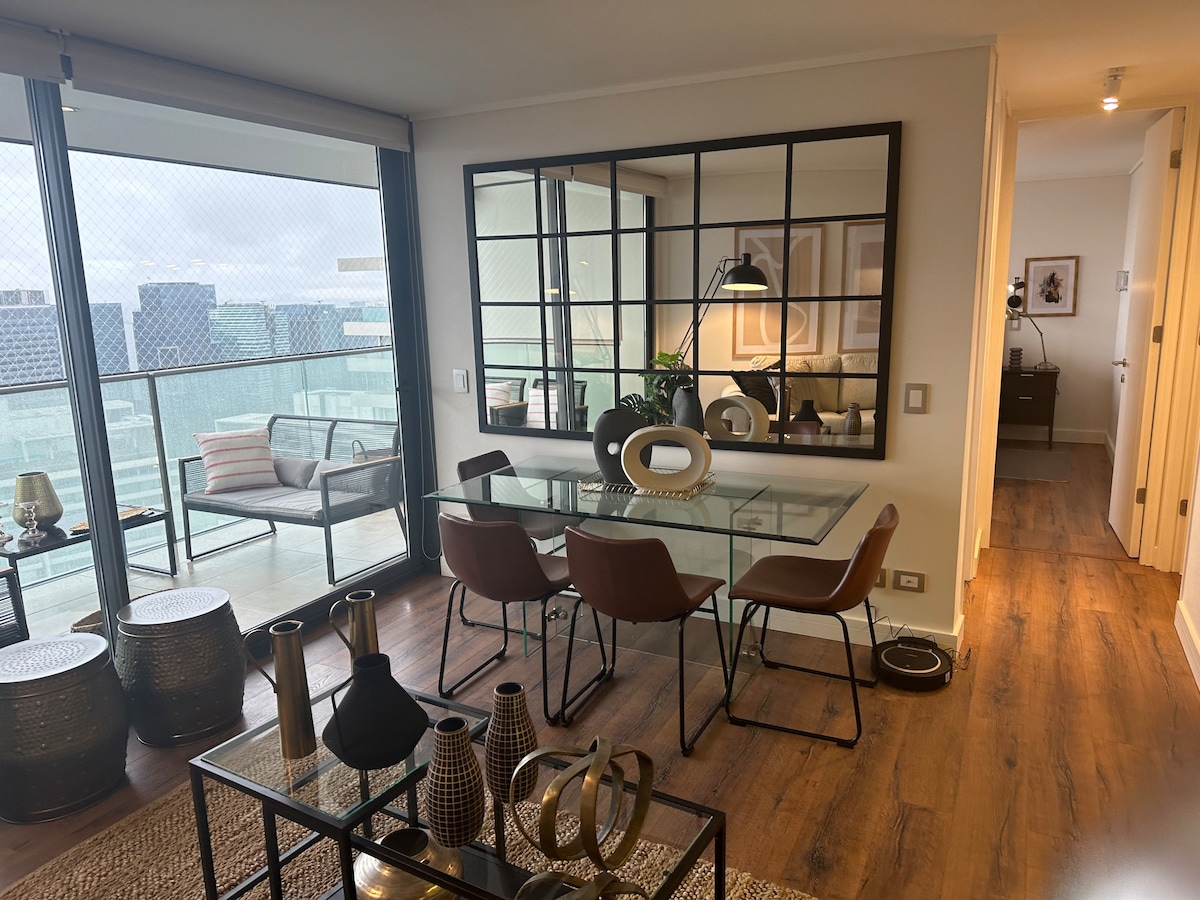 A modern dining area is furnished with a glass table and four brown chairs, accompanied by decorative objects and vases. A large mirror reflects the room, creating a sense of spaciousness. Nearby, a balcony is visible with outdoor seating, framed by large windows.