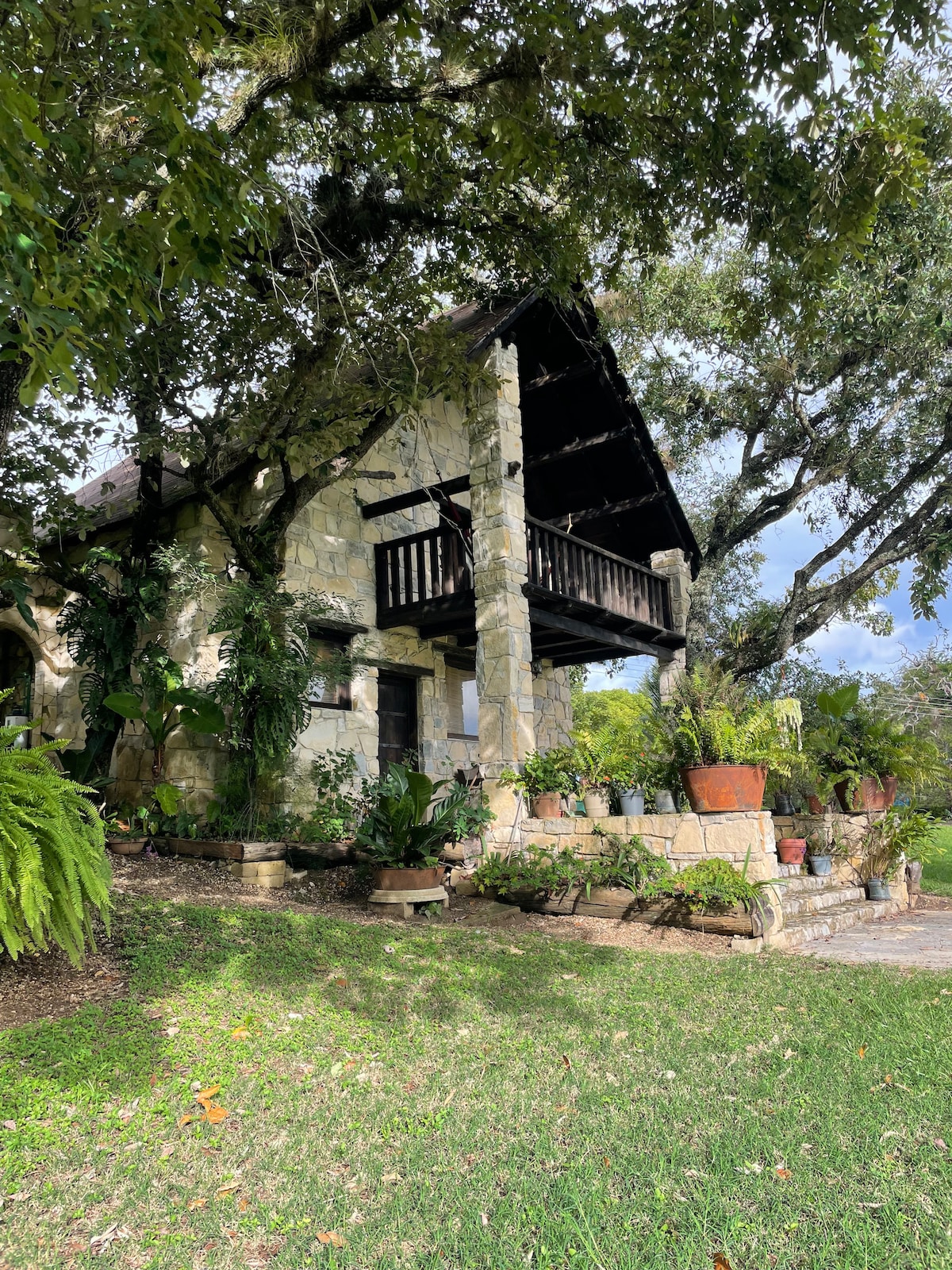 A charming cabin constructed from stone is surrounded by lush greenery. A balcony extends from the upper level, providing a cozy spot to enjoy the view. Potted plants and landscaping details enhance the inviting outdoor space, complemented by ample natural light.