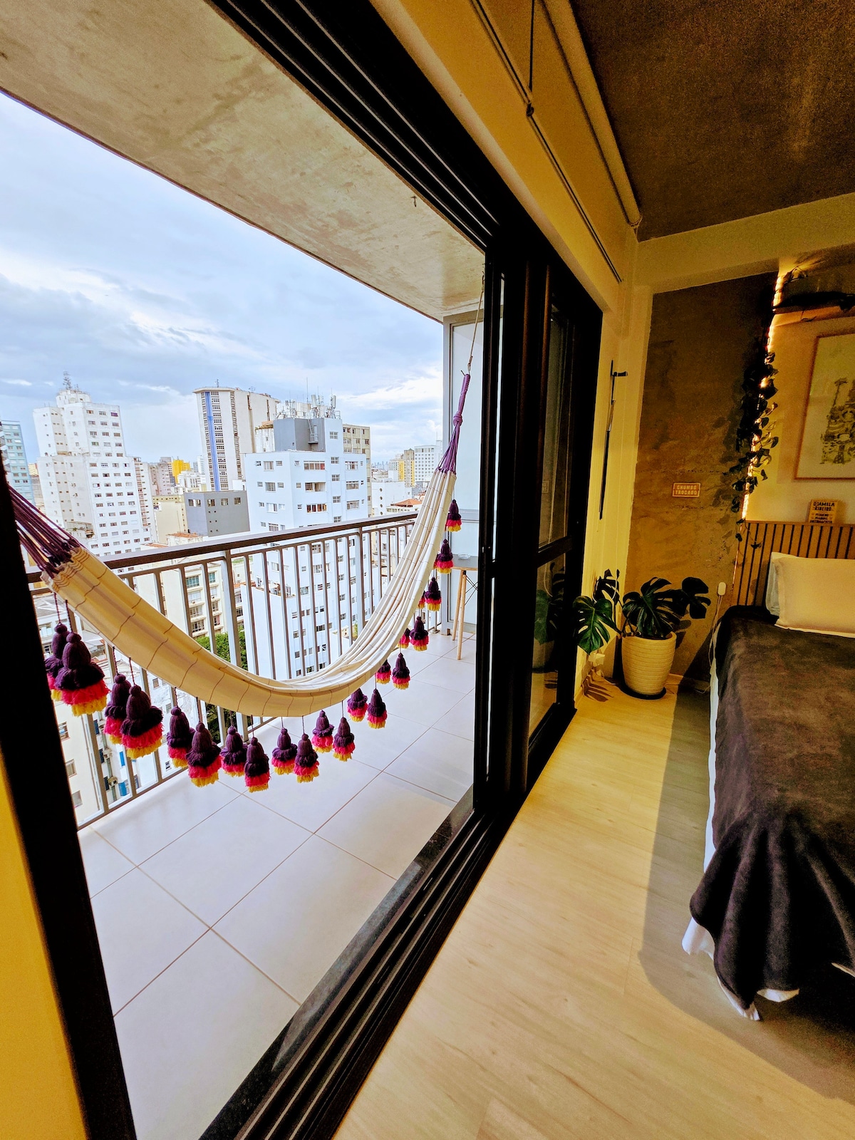 A spacious room features glass doors leading to a private balcony with a colorful hammock. Urban views are visible in the background, showcasing a blend of buildings and a cloudy sky. A plant is positioned nearby, enhancing the serene atmosphere of the space.