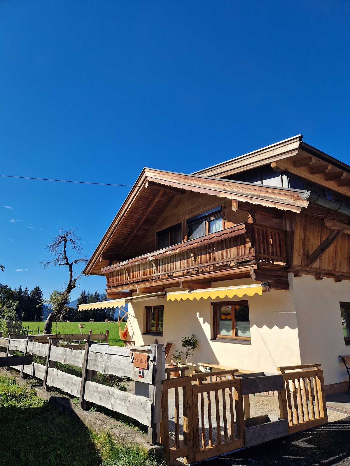 A charming house with a wooden balcony is situated under a clear blue sky. The exterior features natural wood accents and decorative yellow awnings, while a fenced area and greenery surround the property, providing a sense of tranquility. Sunlight enhances the inviting atmosphere.