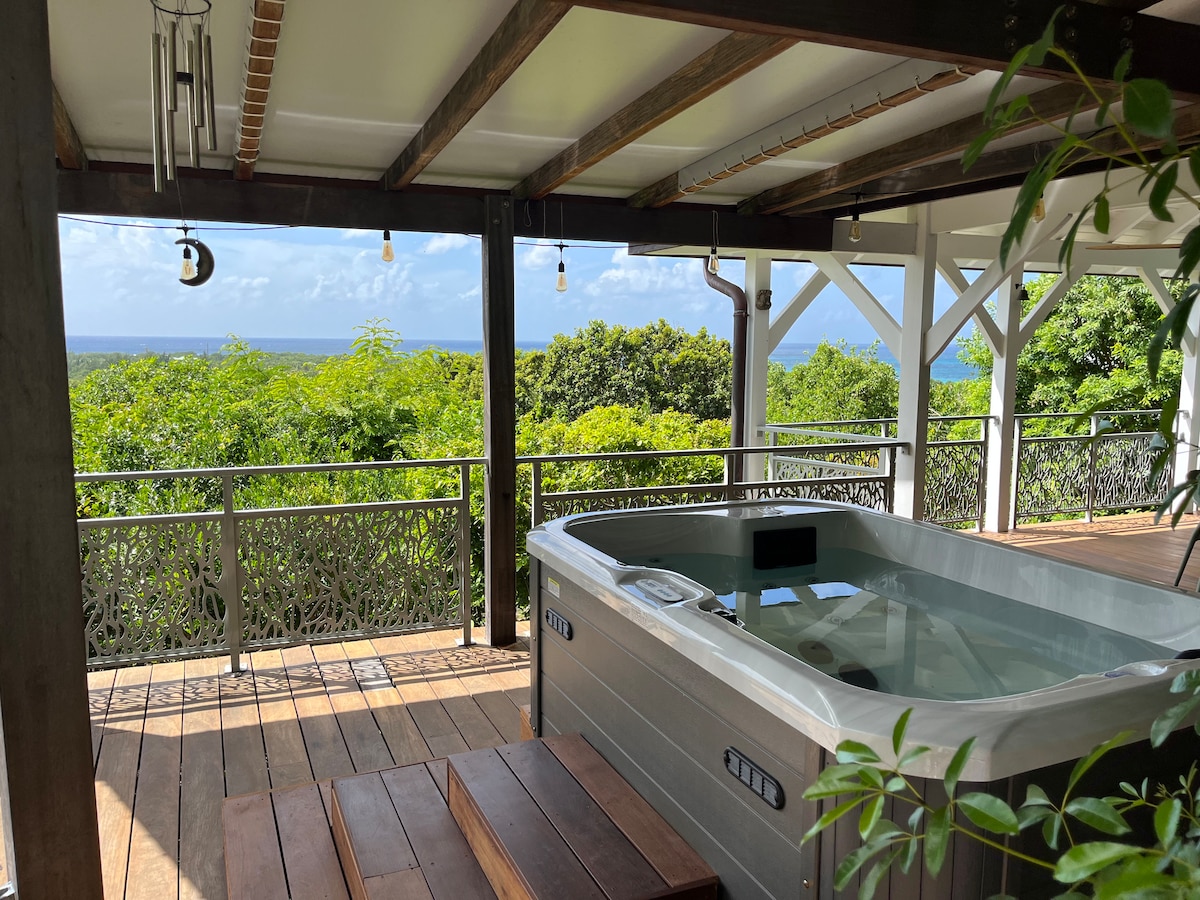 A spacious covered terrace features a hot tub positioned for optimal sea views. The area includes wooden flooring and decorative railings, surrounded by lush greenery. Natural light fills the space, enhancing the serene atmosphere of relaxation and connection with nature.