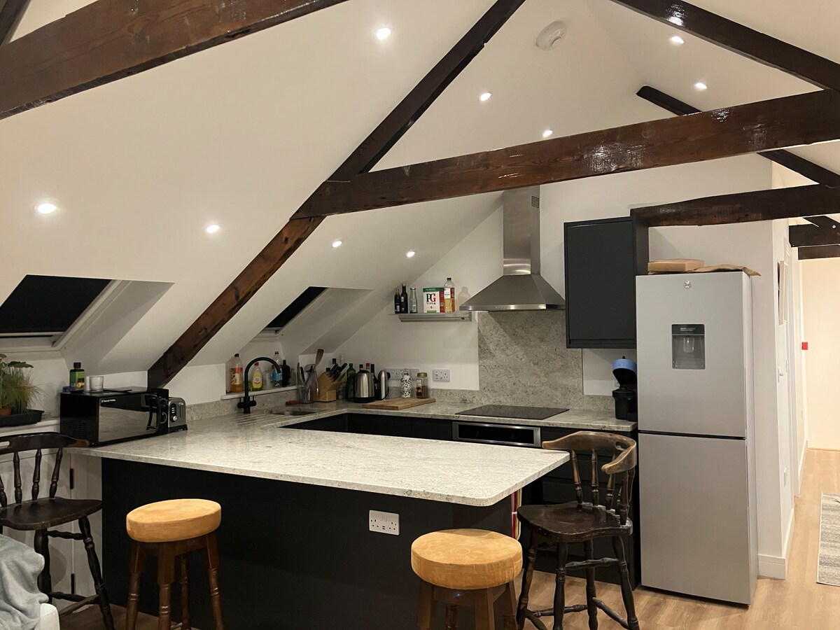 An open kitchen features a large island with seating at three wooden stools. Modern appliances, including a refrigerator and stove, are integrated into the design. Exposed wooden beams create a warm ambiance, complemented by soft overhead lighting and a neutral color palette.