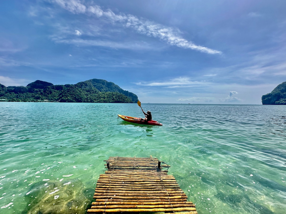 A wooden dock extends into clear turquoise waters, leading to an individual kayaking on a calm day. Lush green hills serve as a backdrop, under a bright blue sky scattered with clouds, creating a serene coastal scene.