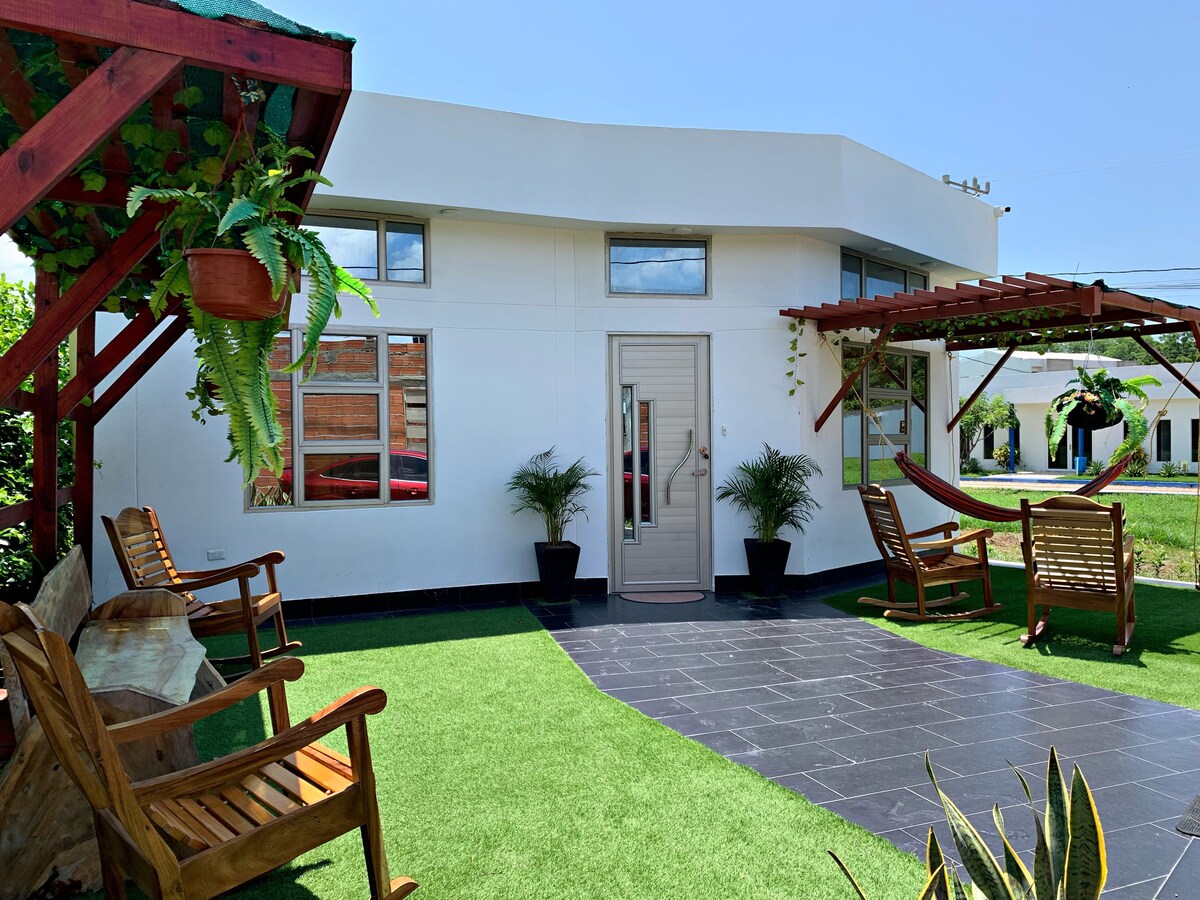 A welcoming entrance features a smooth pathway leading to a light-colored front door. Relaxing seating areas composed of wooden chairs are positioned under a shaded pergola, along with a hammock and greenery complementing the space. The grass-like surface adds a touch of nature.