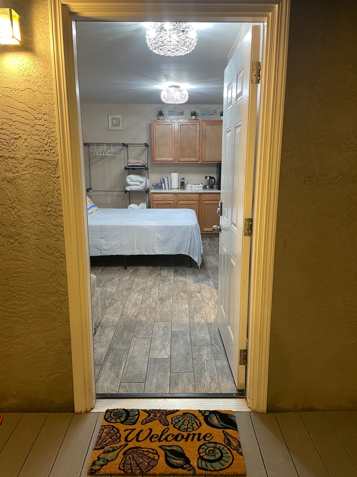 The entrance is framed by a white door, leading into a well-lit room featuring a spacious bed and a cozy kitchenette with wooden cabinetry. A welcome mat with a floral design sits at the threshold, inviting guests inside.