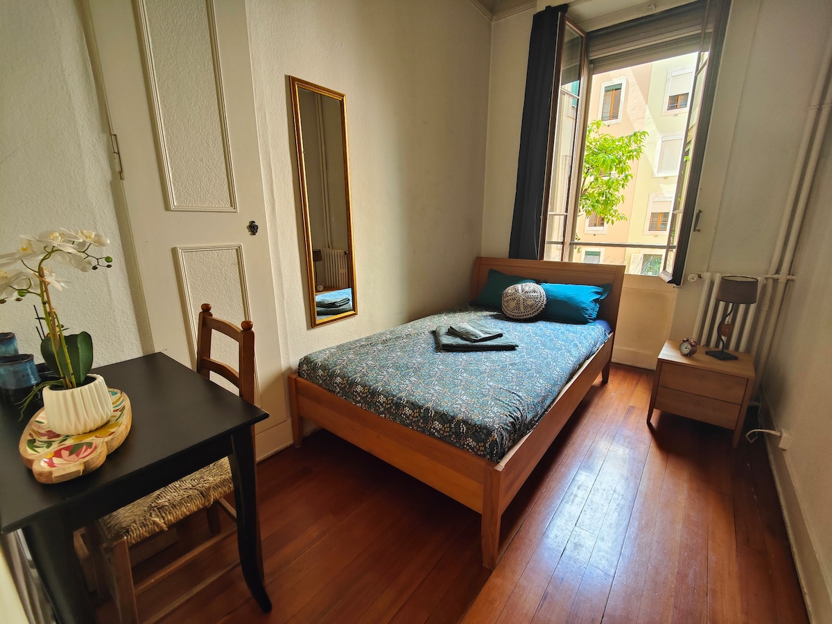 A cozy room features a single bed with a patterned cover and a decorative pillow. A small wooden chair and table are positioned beside the bed. Natural light enters through a window, complementing the room's warm wooden floor and neutral walls. A mirror reflects the space.